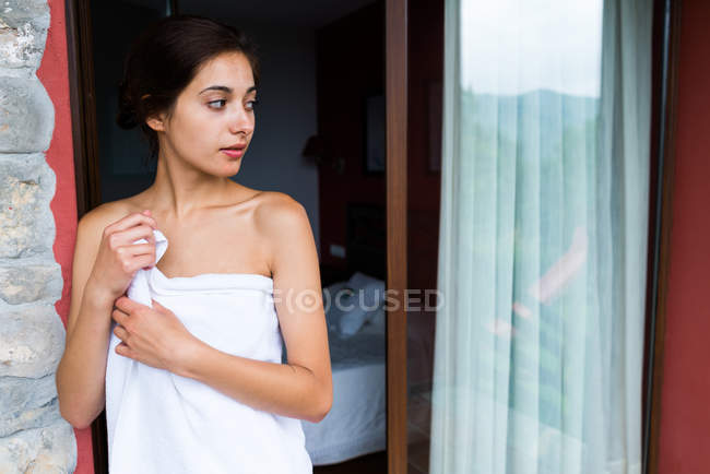 Молодая женщина, завернутая в полотенце, стоит у входа на балкон и смотрит в сторону — стоковое фото