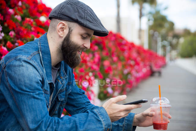 Side view of bearded person sitting and surfing smartphone while drinking smoothie. — Stock Photo