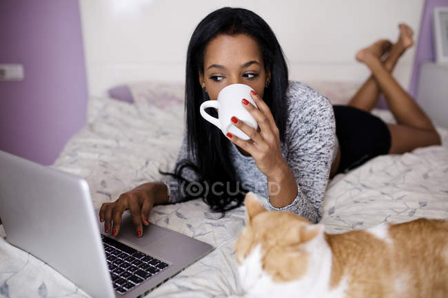 Ragazza che beve caffè mentre naviga laptop a letto — Foto stock