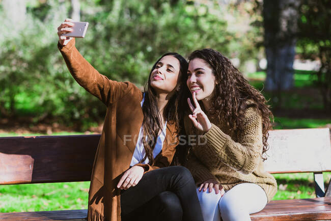 Zwei junge hübsche Freundinnen sitzen auf Bank im Park und machen Selfie. — Stockfoto