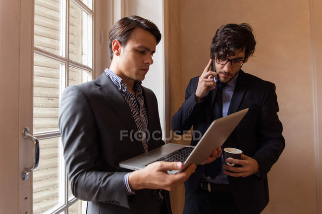Zwei Geschäftsleute in Anzügen stehen mit Laptop am Fenster. — Stockfoto