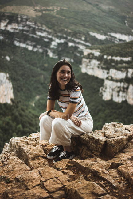 Mujer alegre sentada en piedra y mirando hacia las montañas . - foto de stock