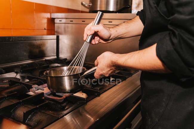 Mid section of chef preparing dish in sauce pot on stove with whisk at restaurant kitchen — Stock Photo