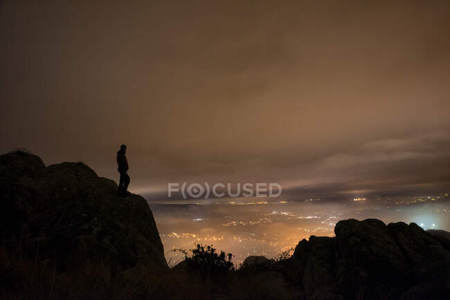 El hombre mira las luces de la ciudad desde la cima de la montaña una fría y nublada noche de invierno - foto de stock
