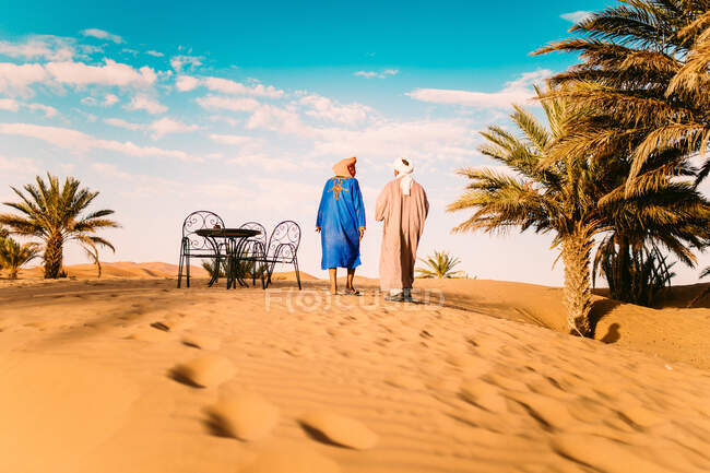 Two people walking near palms in desert. Horizontal outdoors shot — Stock Photo