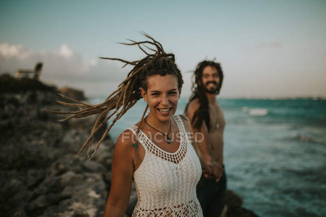 Portrait of happy woman with dreadlocks holding male hand and looking at camera on background of tropical beach. — Stock Photo