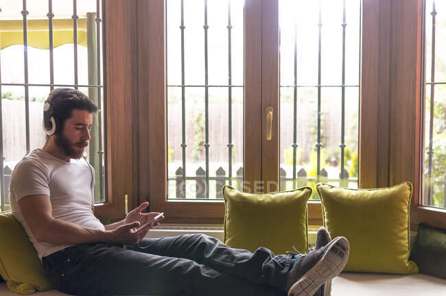 Jeune homme écoutant de la musique avec tablette détendue à la maison avec la fenêtre éclairée par la lumière chaude du soleil — Photo de stock