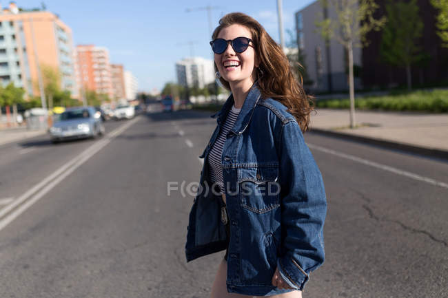 Fröhliche junge Frau mit Sonnenbrille blickt beim Überqueren der Straße in die Kamera. — Stockfoto