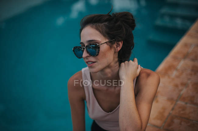 Retrato de mujer morena con estilo en gafas de sol espejadas sentadas en el borde de la piscina - foto de stock