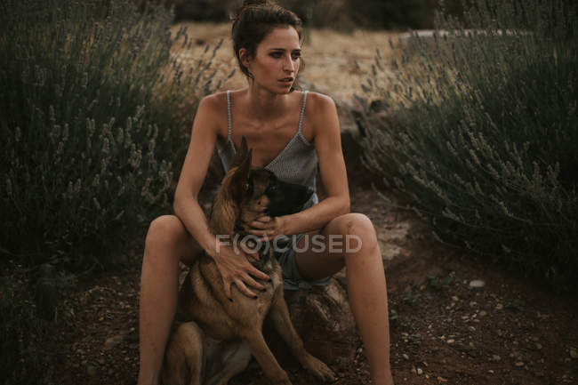 Portrait de brune en robe assise sur la pierre et étreignant chien de berger dans la campagne — Photo de stock