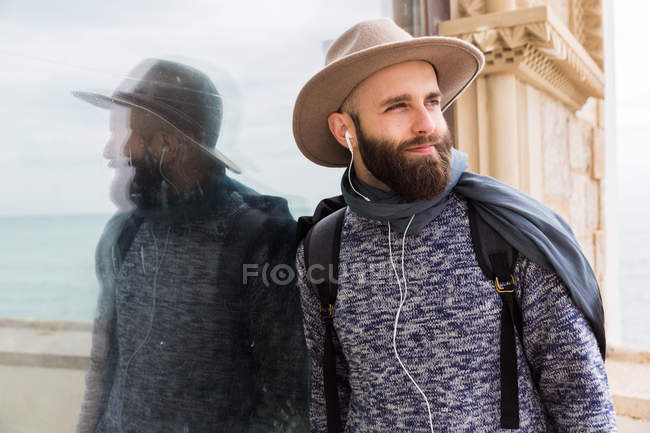 Retrato de hombre barbudo con auriculares apoyados en vidrio y mirando hacia otro lado - foto de stock