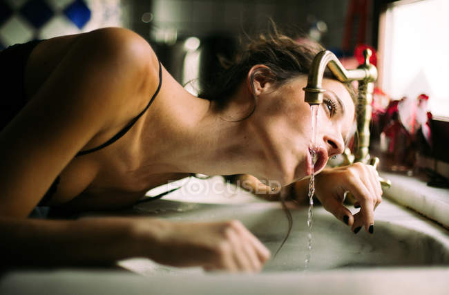Femme assoiffée en lingerie eau potable du robinet vintage à la lumière du jour — Photo de stock