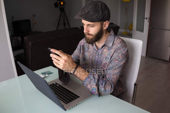 Portrait d'un homme barbu assis à table avec un ordinateur portable et utilisant un smartphone — Photo de stock
