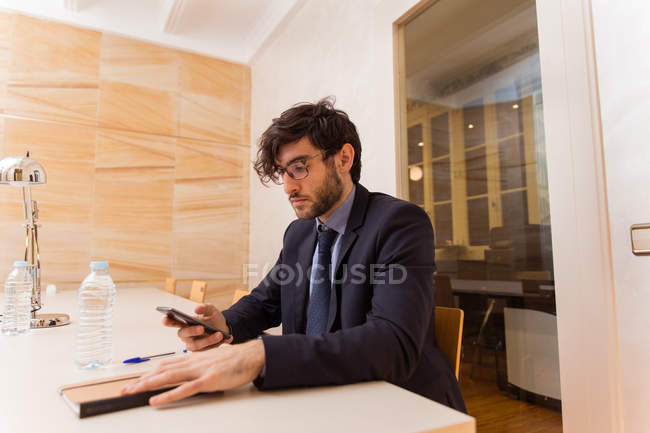 Young businessman in suit sitting in meeting room and browsing smartphone. — Stock Photo