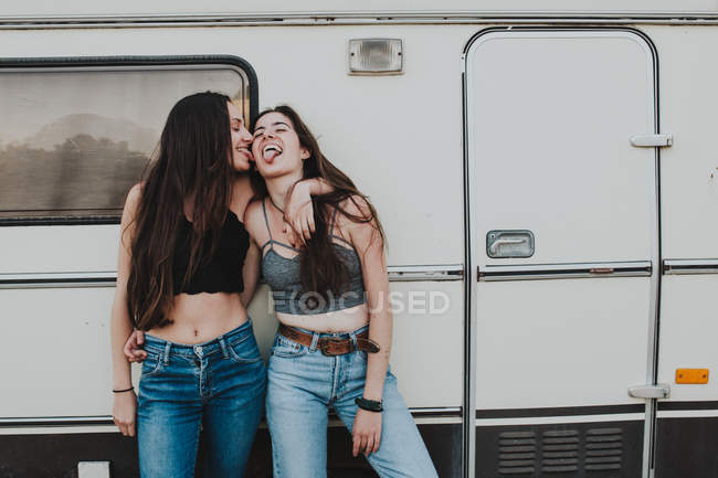 Two cheerful girlfriends hugging and sticking out tongues at trailer — Stock Photo