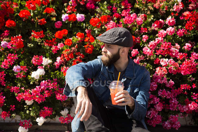 Vorderansicht eines bärtigen Mannes, der neben Blumen sitzt und Smoothie in der Hand hält, während er wegschaut. — Stockfoto
