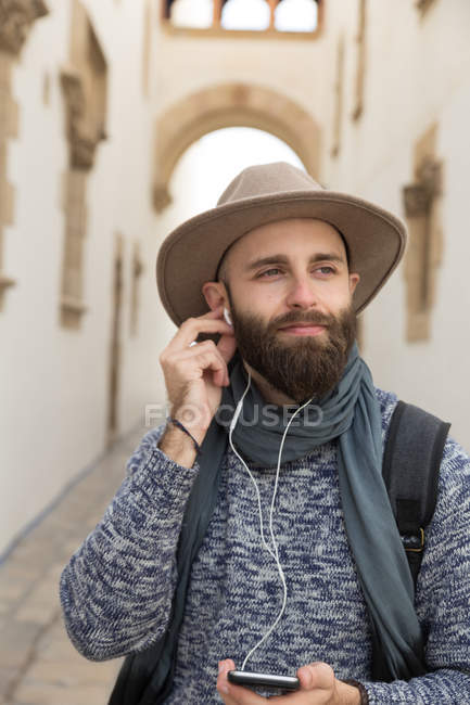 Портрет мечтательного человека с рюкзаком, смотрящего в сторону и надевающего наушники на улице — стоковое фото