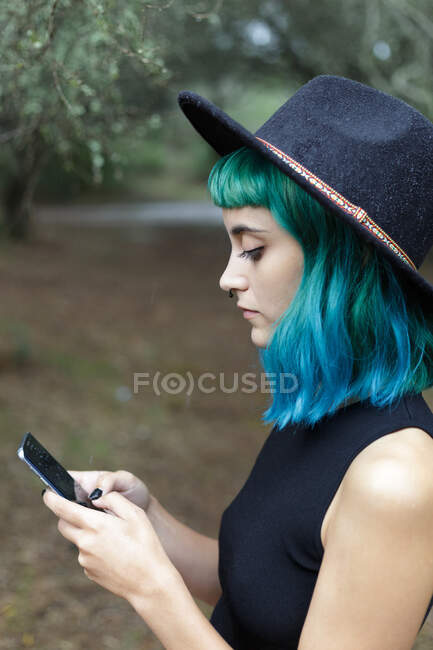 Side view of stylish hipster girl with blue hair in black hat standing outdoor and looking at her cell phone. — Stock Photo