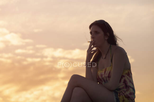 Молода жінка курить цигарку, спостерігаючи за заходом сонця — стокове фото