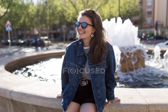 Allegro bella donna seduta e sorridente alla fontana della città . — Foto stock