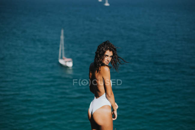 Side view of woman wearing white swimsuit looking to camera and posing against ocean with floating yachts — Stock Photo