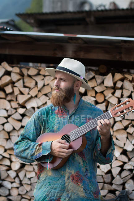 Бородач играет на укулеле над сложенными бревнами. — стоковое фото