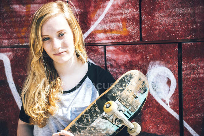 Блондинка-скейтбордистка позирует со скейтбордом у стены граффити — стоковое фото