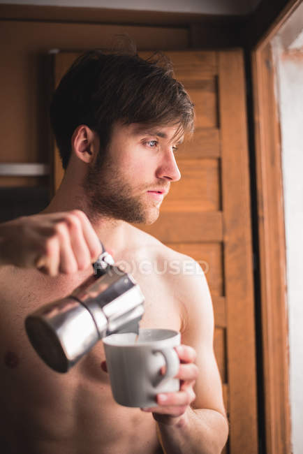 Hemdlos schläfriger Mann, der Kaffee in Becher gießt und zum Fenster schaut. — Stockfoto