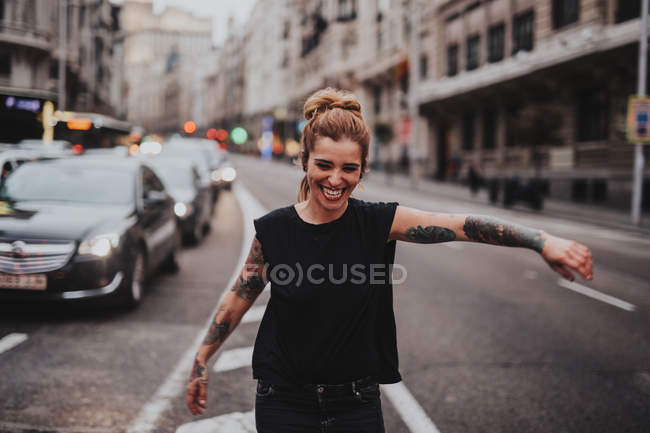 Retrato de chica alegre con brazos tatuados bailando en medio de la carretera con el tráfico - foto de stock