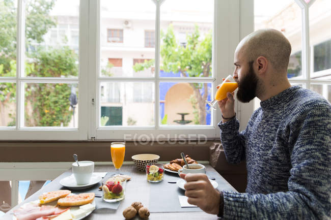 Horizontal en interiores disparo de hombre tomando jugo de naranja fresco desayunando en la cafetería . - foto de stock