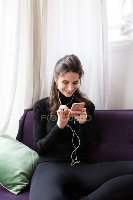 Smiling brunette woman in earphones sitting on couch and browsing smartphone — Stock Photo