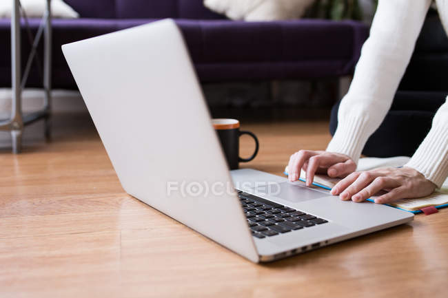 Ritaglia l'immagine della digitazione femminile sul computer portatile sul pavimento con notebook e tazza — Foto stock