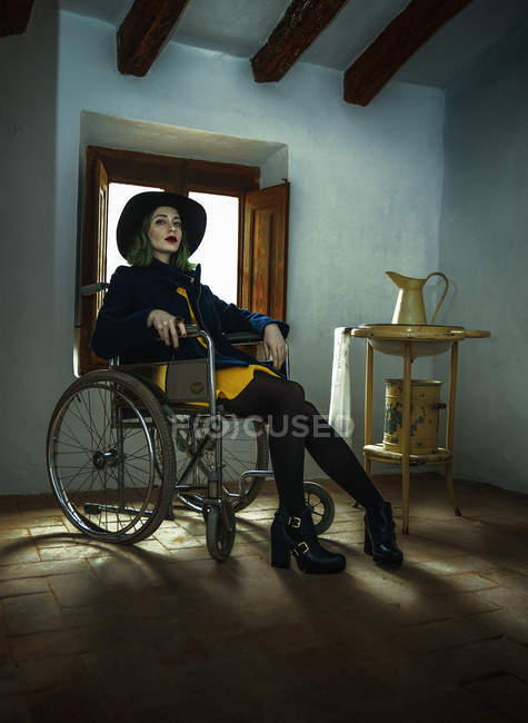 Молодая женщина сидит в инвалидной коляске и смотрит в камеру напротив окна — стоковое фото