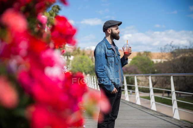 Vista laterale dell'uomo barbuto in piedi con tazza di frullato vicino a fiori luminosi . — Foto stock