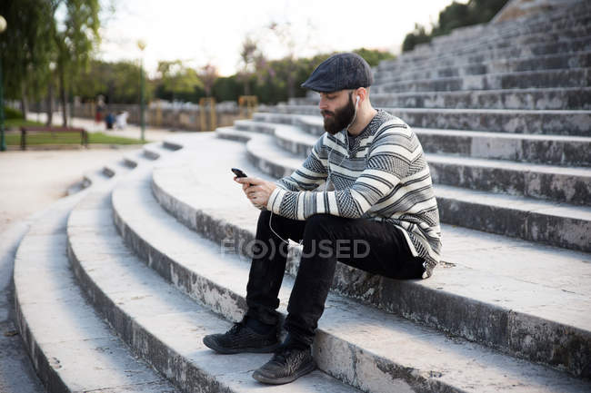 Vista lateral del hombre barbudo sentado en las escaleras y escuchando música - foto de stock