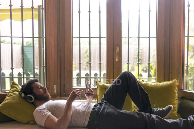 Jovem ouvindo música com tablet relaxado em casa com a janela iluminada pela luz quente do sol — Fotografia de Stock