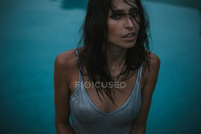 Retrato de morena sensual com cabelo molhado em pé na piscina e olhando para a câmera — Fotografia de Stock