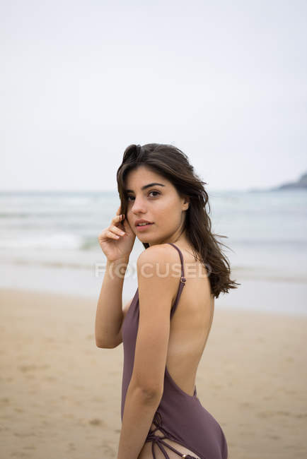 Brunette girl in swimsuit posing on beach and looking over shoulder at camera — Stock Photo