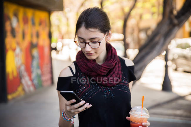 Jeune femme à la rue et avoir smoothie tout en naviguant sur smartphone . — Photo de stock