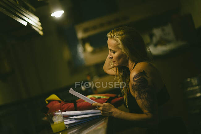 Side view portrait of senior tattooed woman sitting at bar with glass of juice reading papers. — Stock Photo