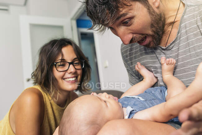 Close-up de adulto feliz pai barbudo brincando com seu bebê em pernas ao lado de mãe sorridente — Fotografia de Stock