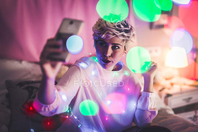 Junge Frau im Märchenlicht schießt auf ihrem Smartphone auf sich selbst. Horizontale Innenaufnahmen. — Stockfoto