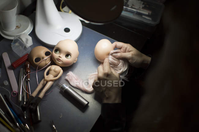 Sobre a vista do ombro das mãos do artista aplicando o cabelo na cabeça da boneca na mesa — Fotografia de Stock