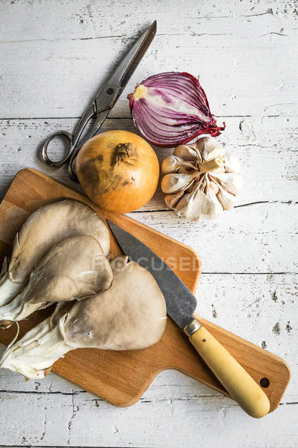 Top view of board with pleurotus mushrooms and rural knife on rustic table with onion and garlic — Stock Photo