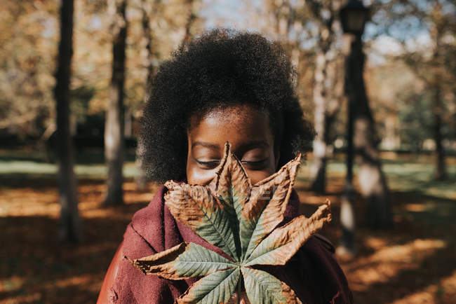 Portrait of girl hiding face with autumn chestnut leaf at park on sunny day — Stock Photo