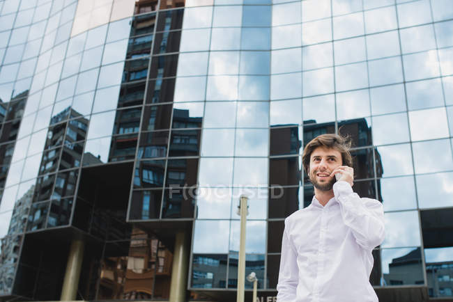 Портрет бізнесмена в білій сорочці, що говорить на смартфоні над фасадом бізнес-будівель на фоні — стокове фото