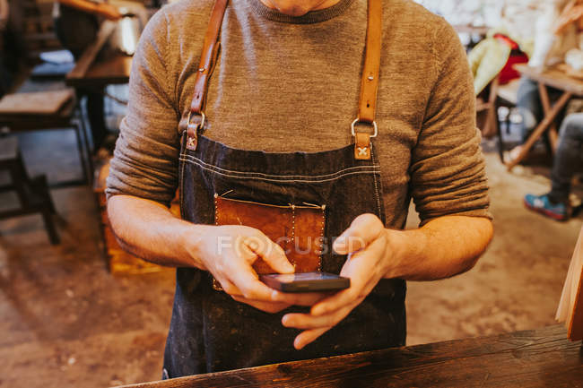 Середня частина чоловіка, одягнена в джинсовий фартух за допомогою смартфона . — стокове фото
