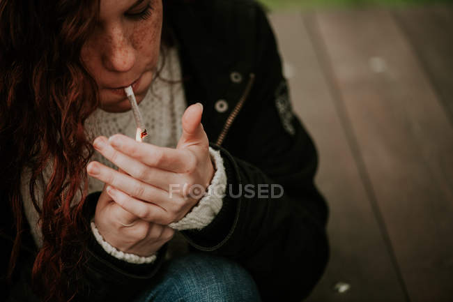 Рыжая девушка с веснушками сидит и закуривает сигарету — стоковое фото