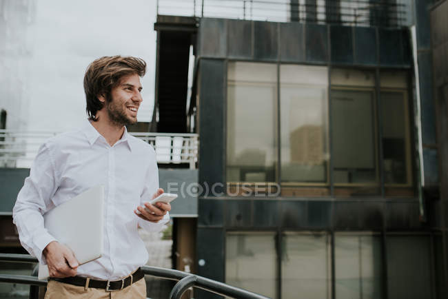 Portrait of bearded businessman holding laptop and cell while smiling away — Stock Photo
