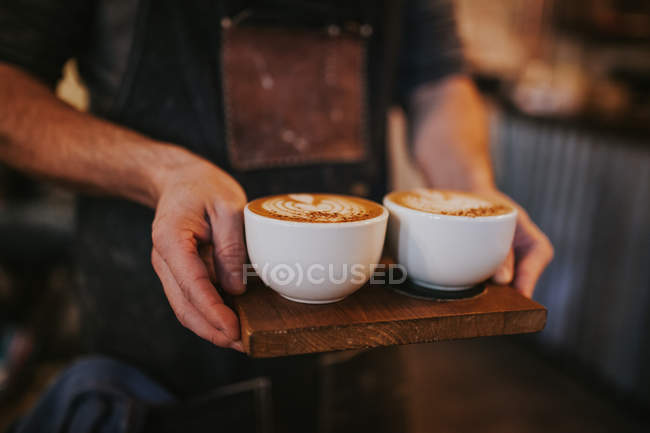 Crop man holding wooden board with two mugs ofcappuccino — Stock Photo
